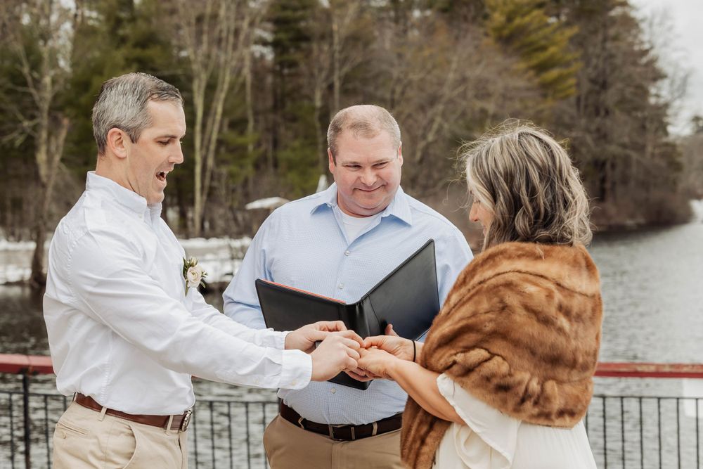 An officiant conducts a wedding ceremony for a couple on a deck overlooking a lake.