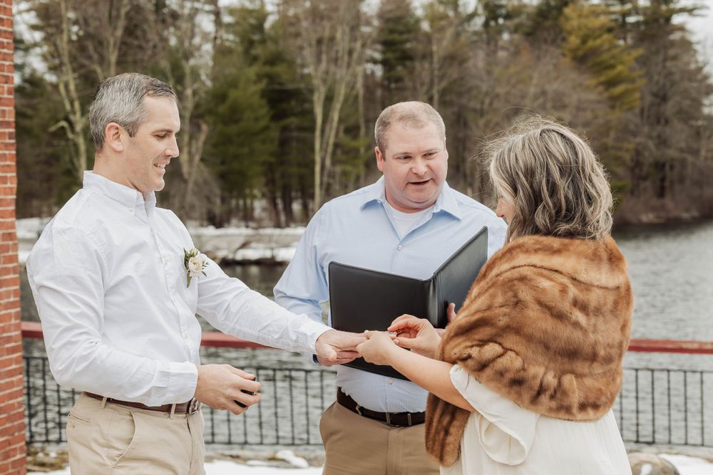 A wedding ceremony takes place on a snowy deck overlooking a lake as a couple exchanges vows with an officiant.