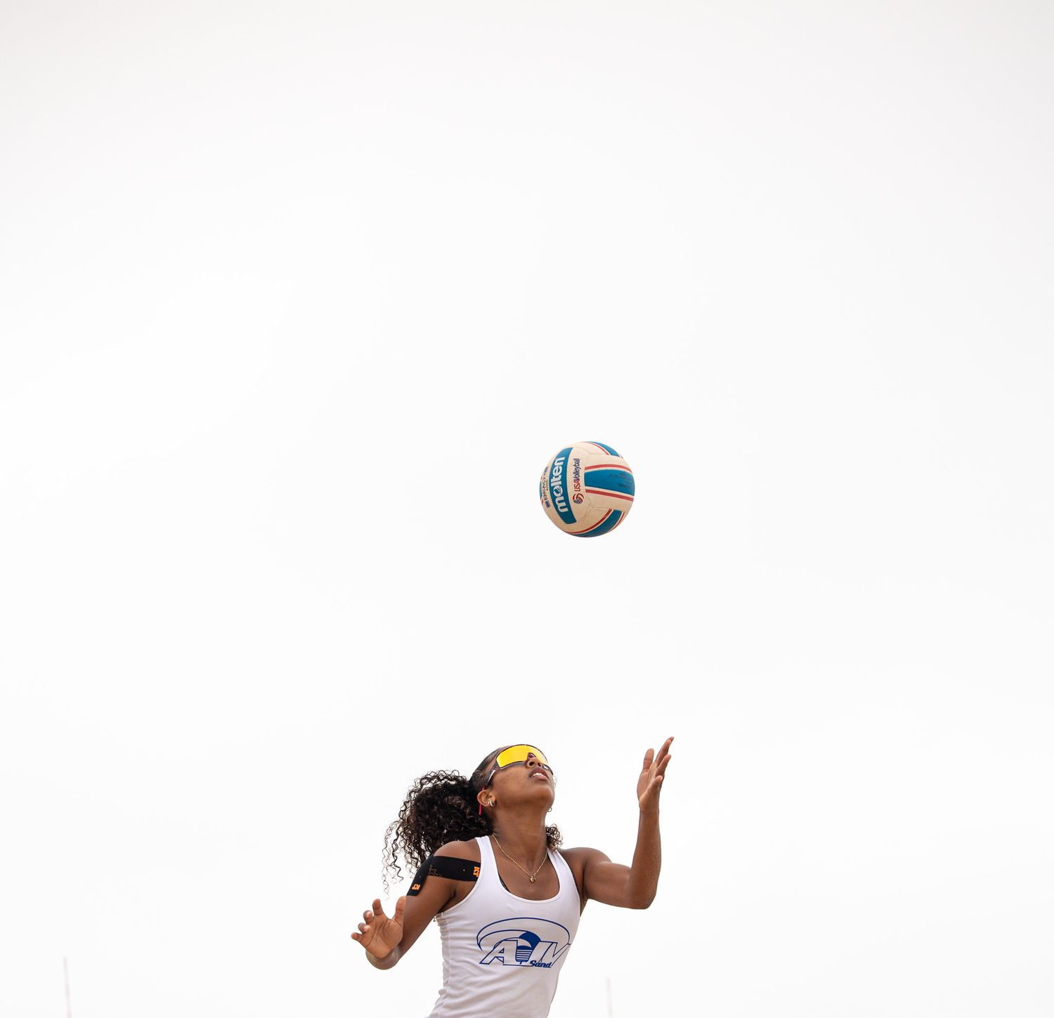 Beach volleyball player jumps to serve the ball against a white sky background.