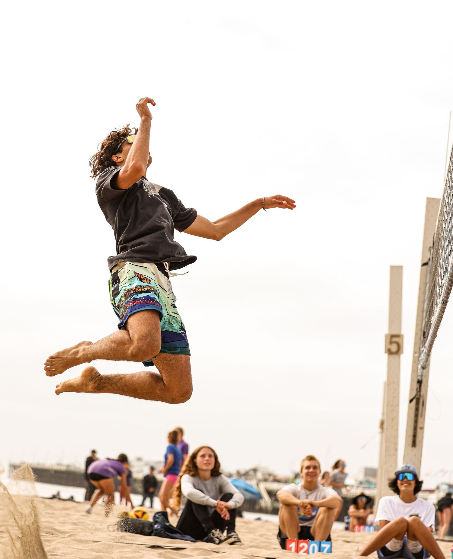 Player leaps high in the air during beach volleyball action with spectators watching.