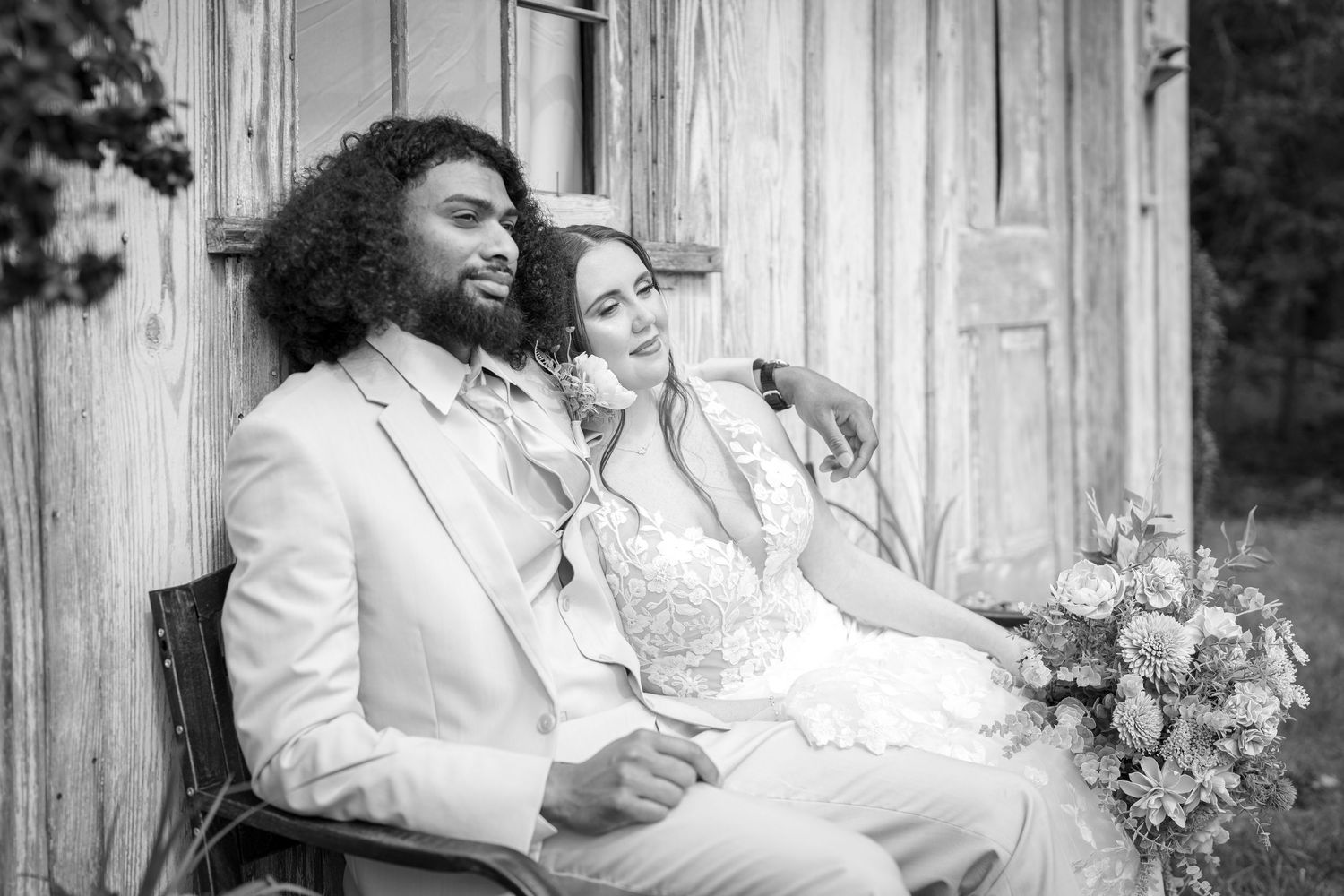 Two people in wedding attire share a joyful moment while sitting together on a bench with flowers beside them.