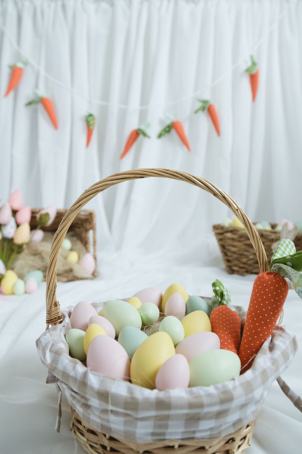 Easter basket filled with pastel colored eggs and decorative carrots, with carrot garland hanging in background.