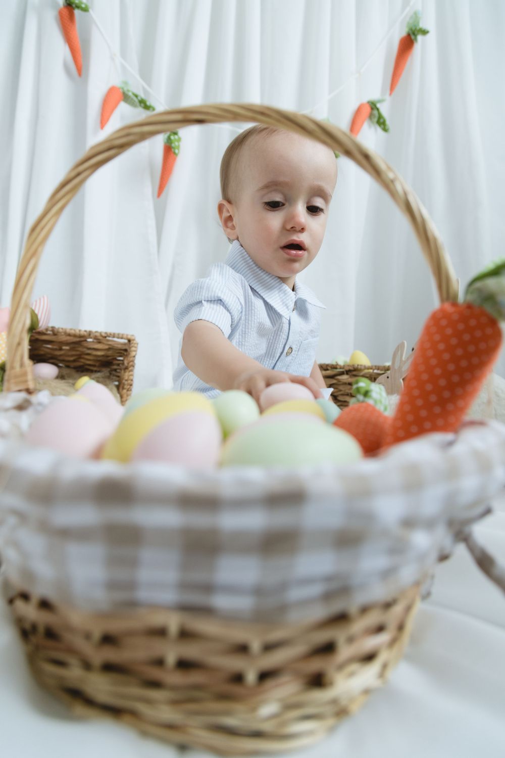 Easter themed photo setup with wicker basket filled with pastel eggs and decorative carrot garland against white backdrop.
