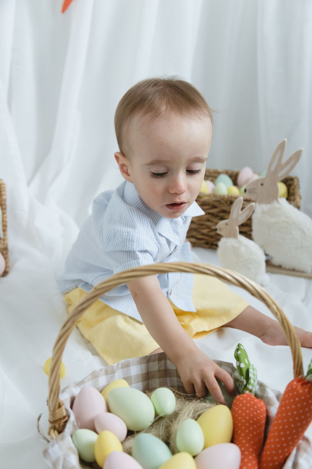 Easter themed photo with wicker basket full of pastel colored eggs and decorative plush bunny against white backdrop.