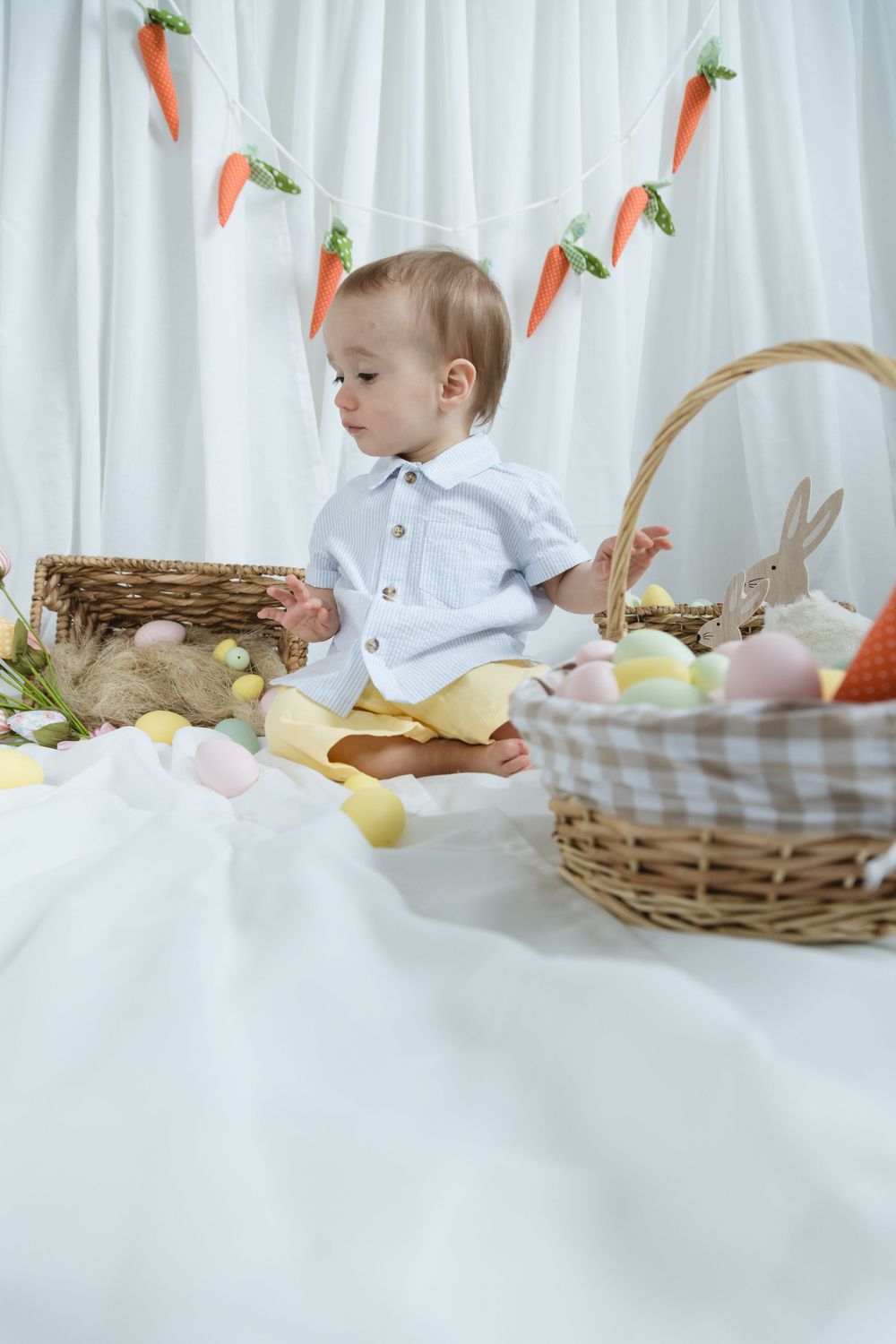 A sweet Easter setup with pastel colored eggs, wicker baskets, and decorative carrot garland against a white curtain backdrop.