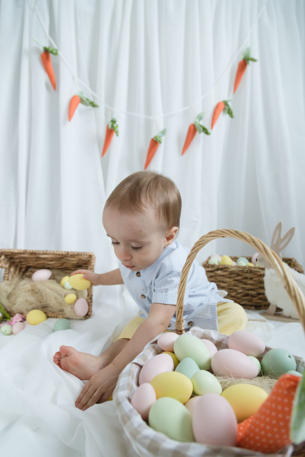 Easter setting with pastel colored eggs, carrot garland decorations, and wicker baskets on white backdrop.