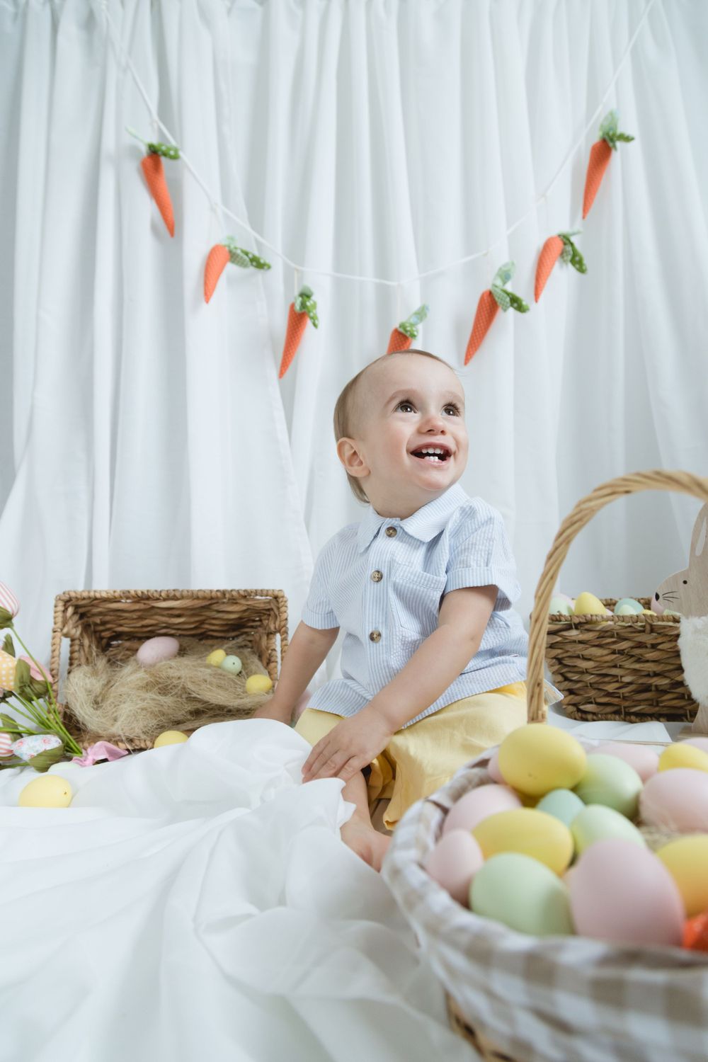 Sweet spring scene with joyful baby surrounded by Easter decor including carrot garland, baskets and pastel eggs on white.