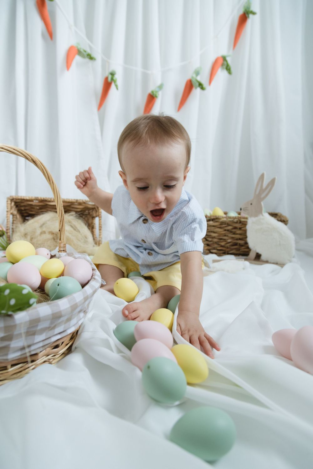 Festive Easter scene with colorful eggs, wicker baskets, bunny decorations and carrot banner on white fabric.