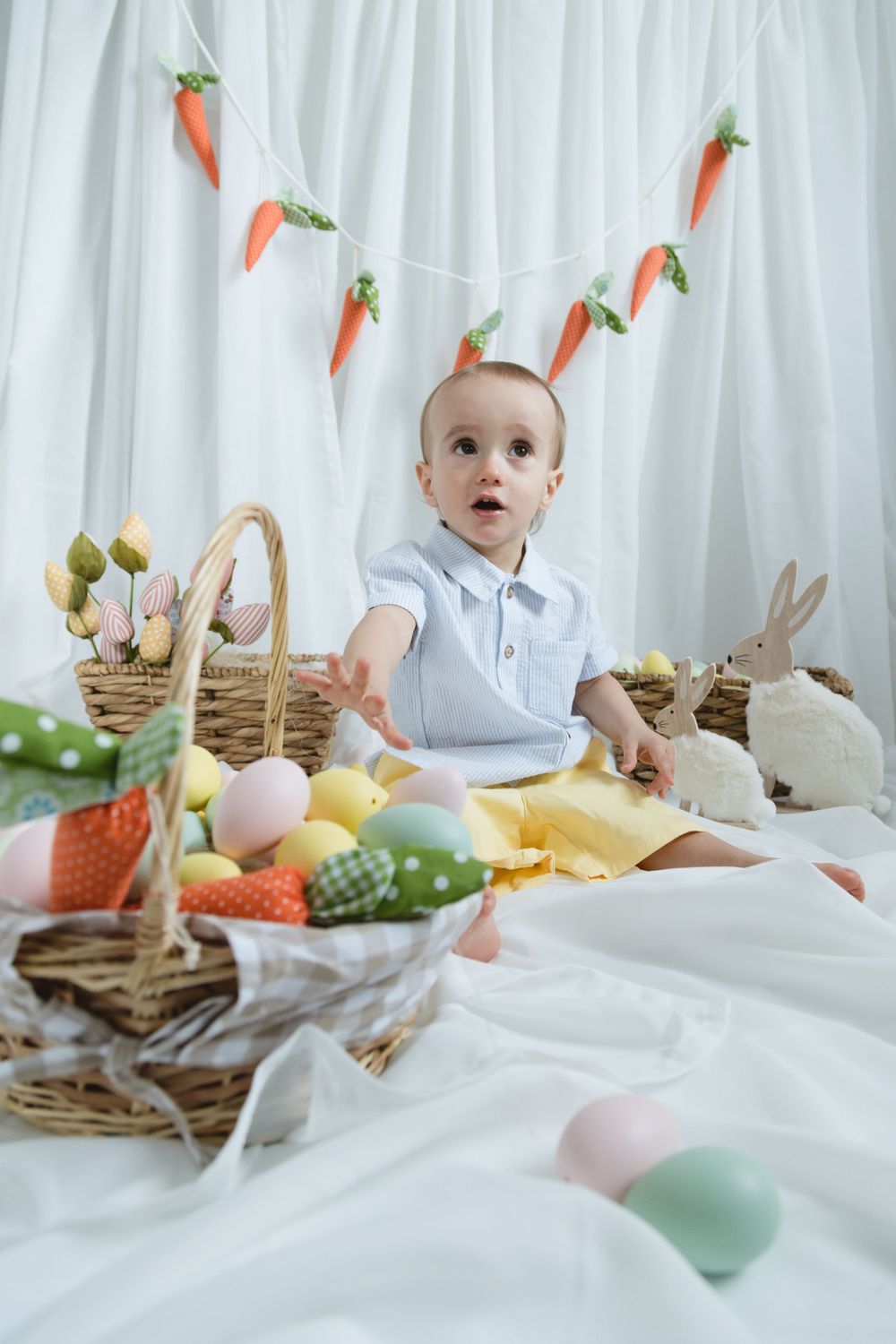 Easter setup with a baby on white backdrop surrounded by baskets, decorative carrots, bunnies and pastel colored eggs.
