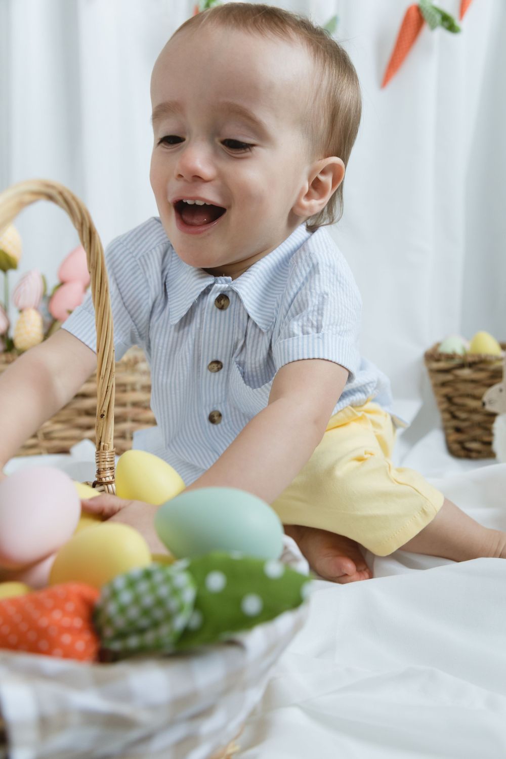 A young child in a striped shirt reaches for colorful Easter eggs in a basket during a festive holiday photoshoot.