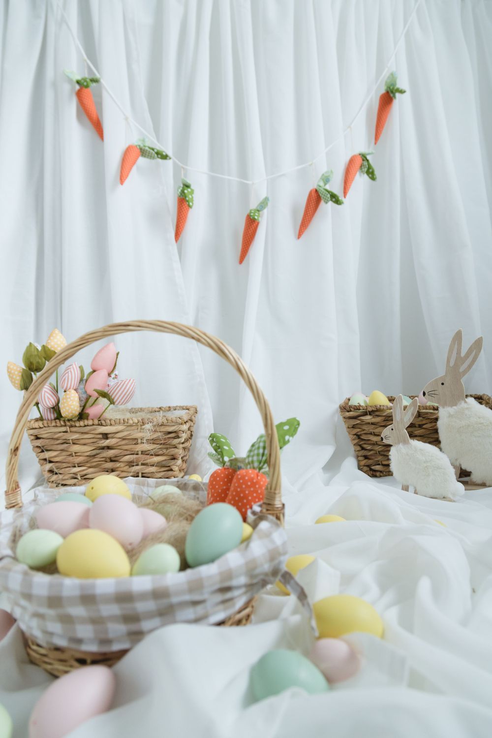 Easter setup with carrot garland, wicker baskets filled with pastel eggs and bunny decorations against white backdrop.