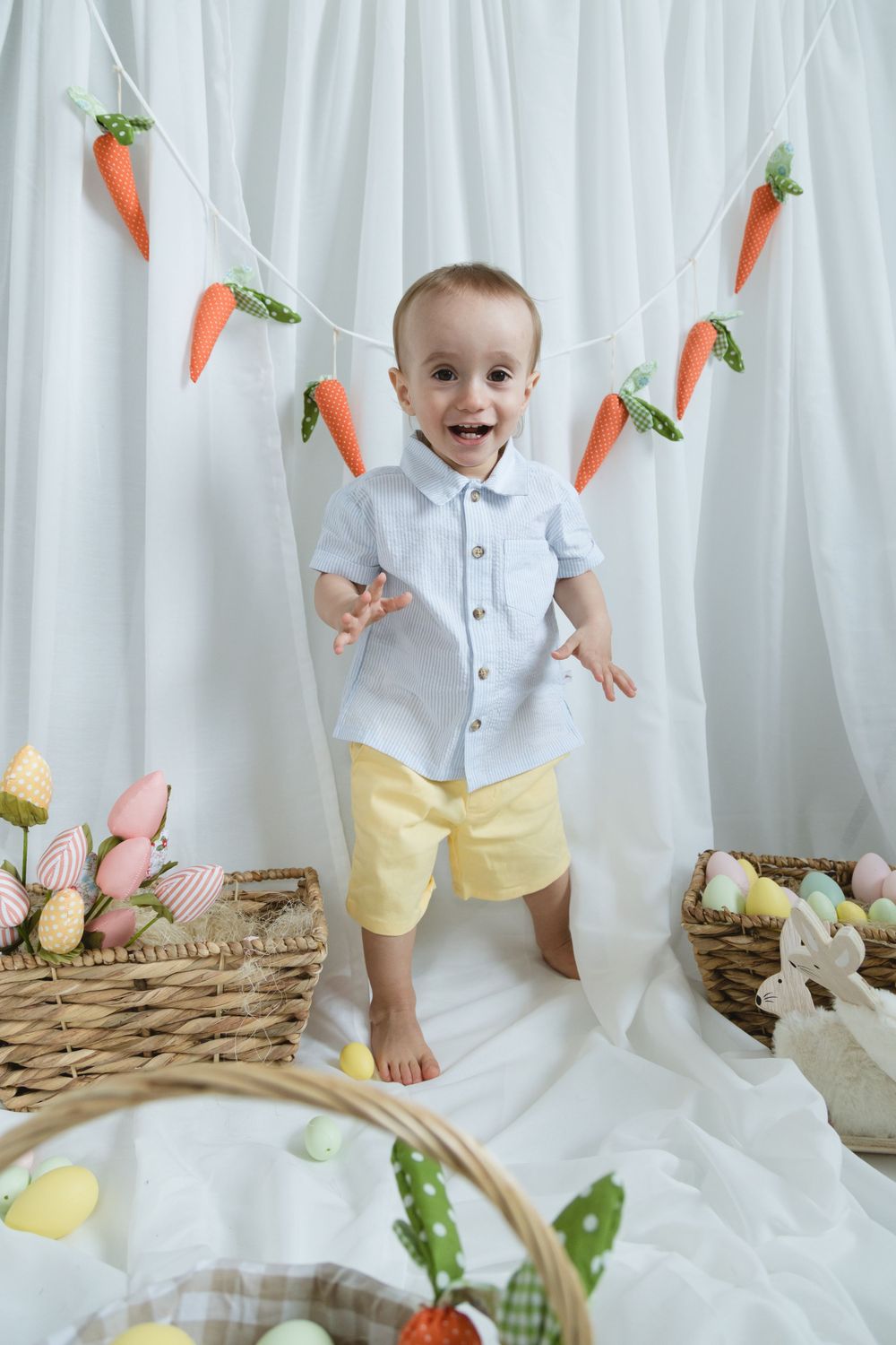 Spring themed photo setup with Easter baskets, pastel eggs, and carrot garland against white curtain backdrop.