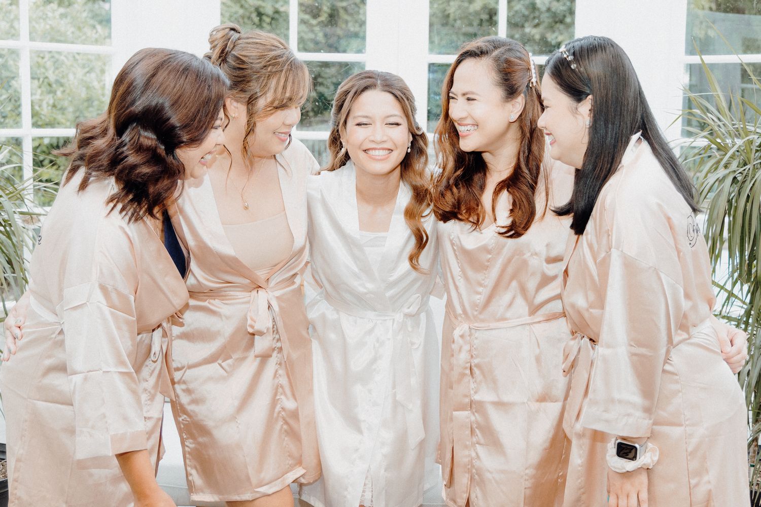 Bridal Party wearing matching blush pink satin robes laughing together captured by Apertured Media Auckland Wedding Photographer.