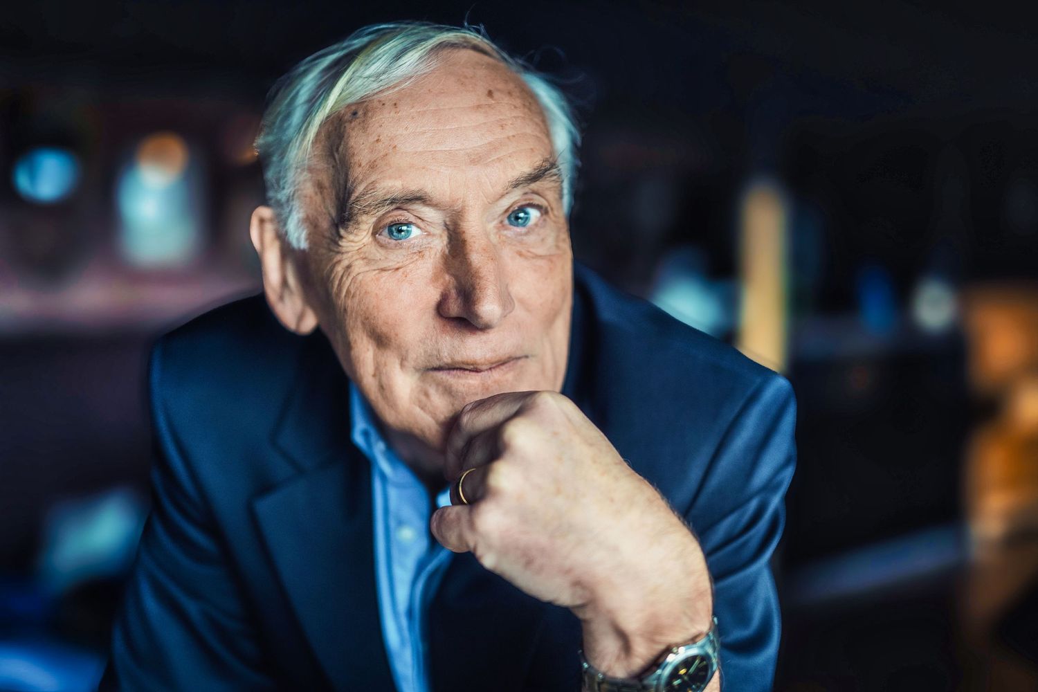Professional man in navy suit poses with hand on chin against dark background.