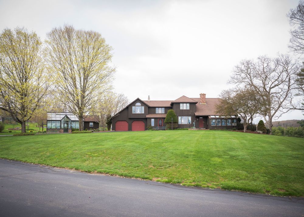 Brick red country house with dormer windows sits on expansive green lawn surrounded by spring trees.
