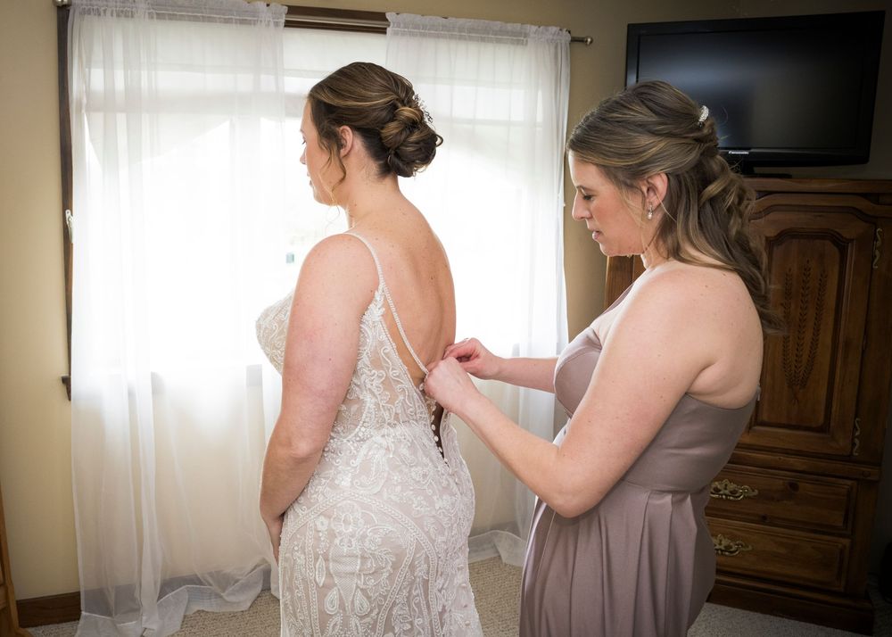 A woman in a lavender bridesmaid dress helps adjust the lacing on a white lace wedding gown in a sunlit room.