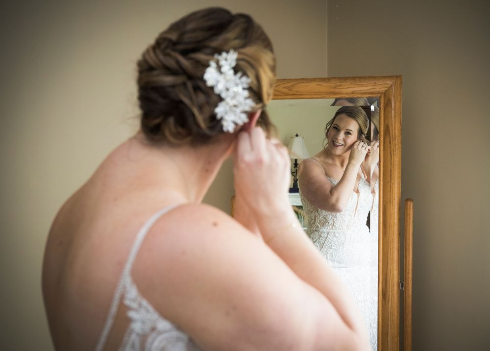 Bride in white lace gown adjusts floral hair accessory while looking in vintage-framed mirror.