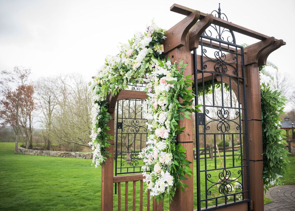 A rustic wooden wedding arch adorned with white flowers and greenery stands in a grassy field.