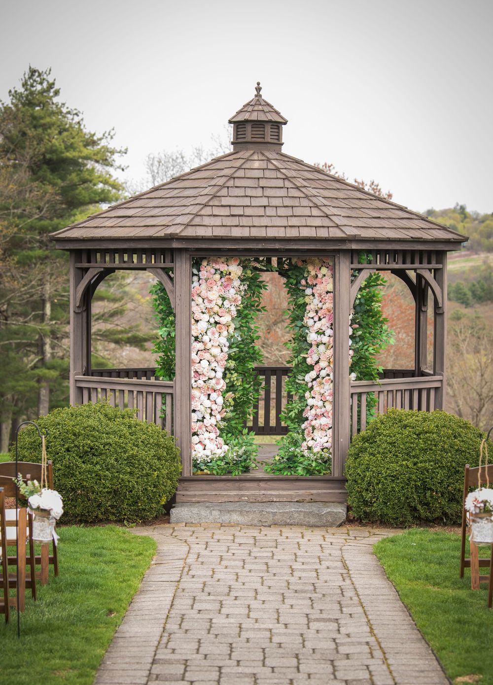 A wooden gazebo decorated with floral arrangements and greenery along a stone pathway for an outdoor wedding ceremony.