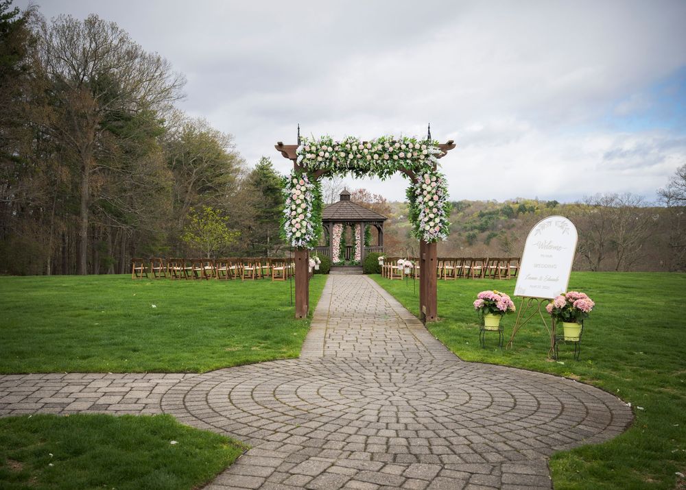 Outdoor wedding ceremony setup with floral arch and stone pathway leading to rustic wooden gazebo in a grassy field.