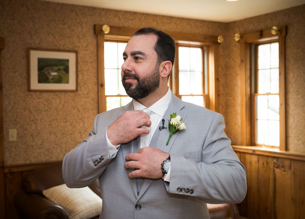 A groom in a light grey suit adjusts his tie while getting ready in a room with windows and wooden walls.