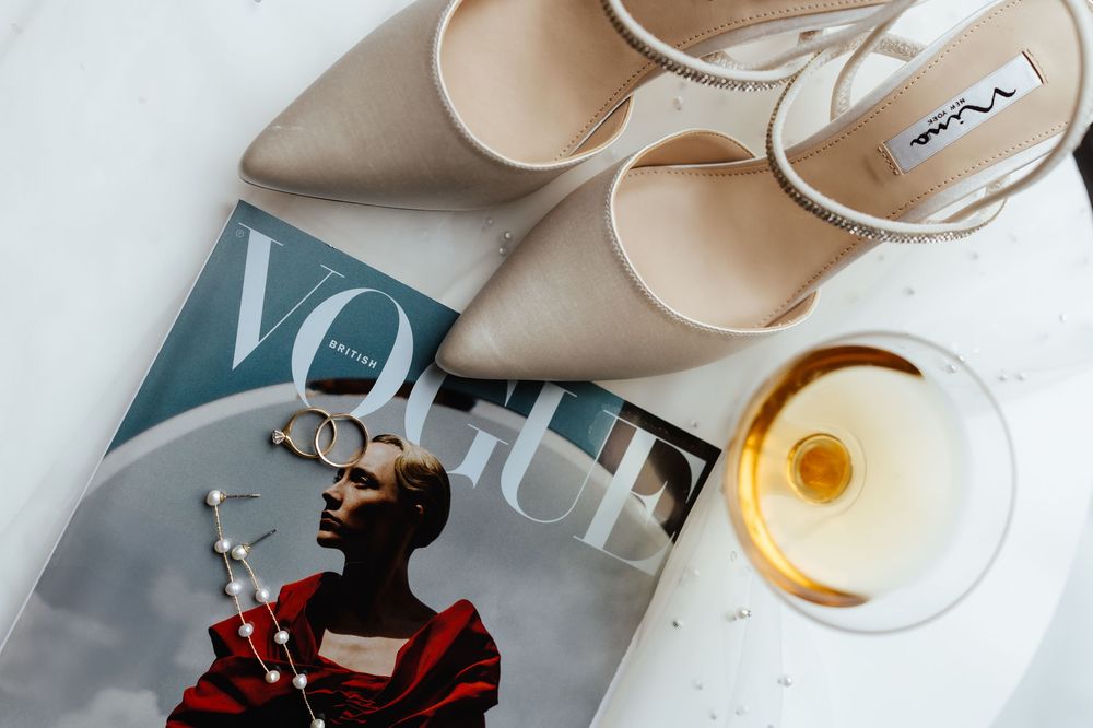 Wedding accessories including nude heels, wedding rings, and a Vogue magazine on white surface.