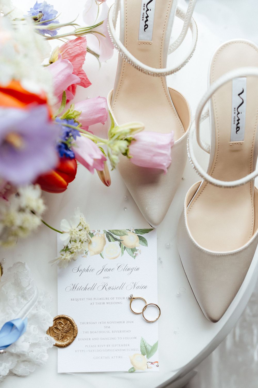Elegant white bridal sandals and wedding rings displayed with soft pink flower petals and invitation cards on marble surface.