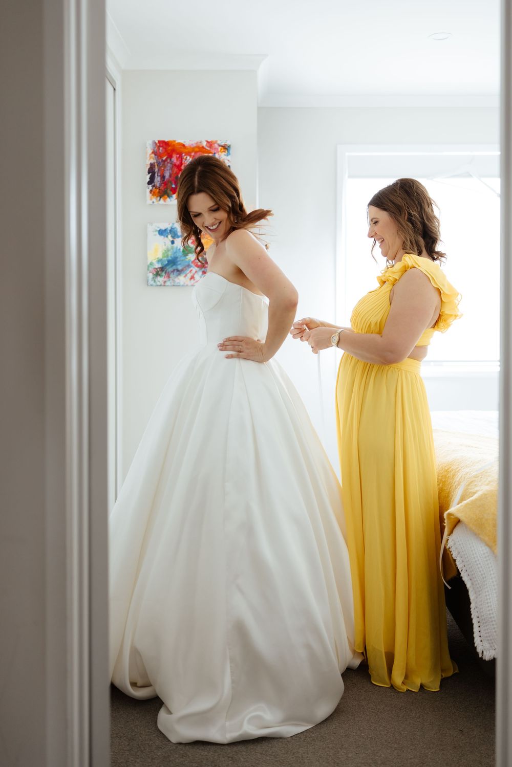 A person in a white wedding gown twirls joyfully while another in a yellow bridesmaid dress looks on in a bright room.