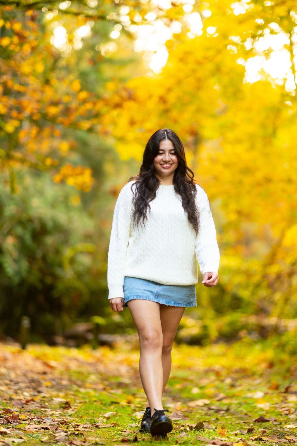Someone wearing a white sweater and denim shorts poses playfully in a sunlit fall forest setting with golden autumn foliage.