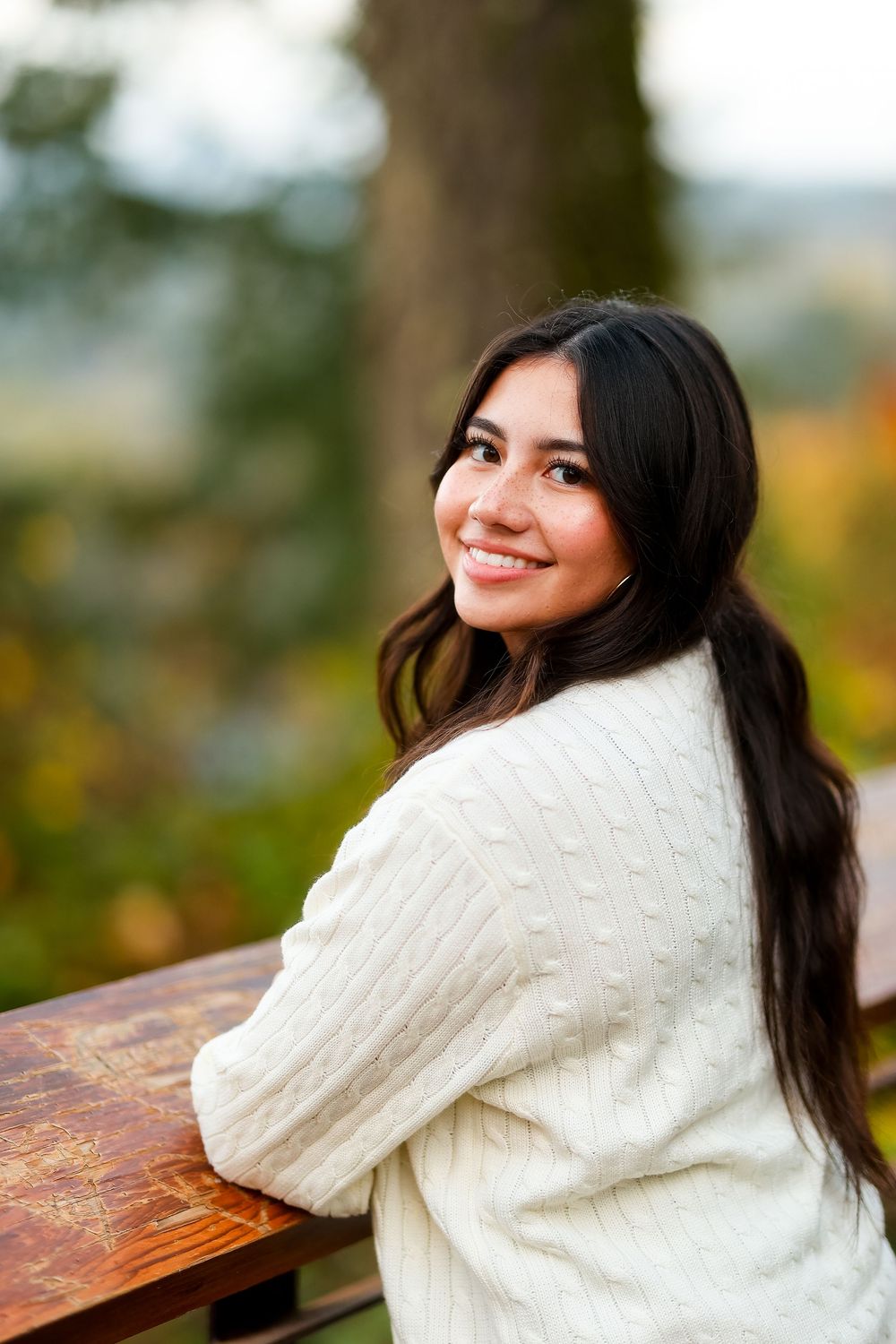 A person in a cozy white cable knit sweater sits outdoors and smiles while looking over their shoulder.