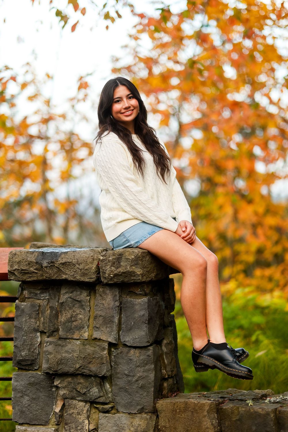 A person in a white sweater and denim shorts sits on a stone wall with autumn foliage in the background.