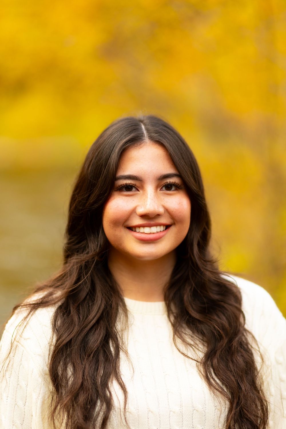 A person wearing a white sweater smiles warmly against a vibrant autumn background with golden yellow foliage.