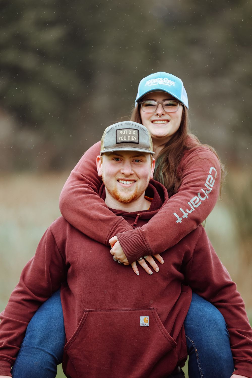 Couple in matching burgundy Carhartt hoodies share a playful piggyback moment outdoors.