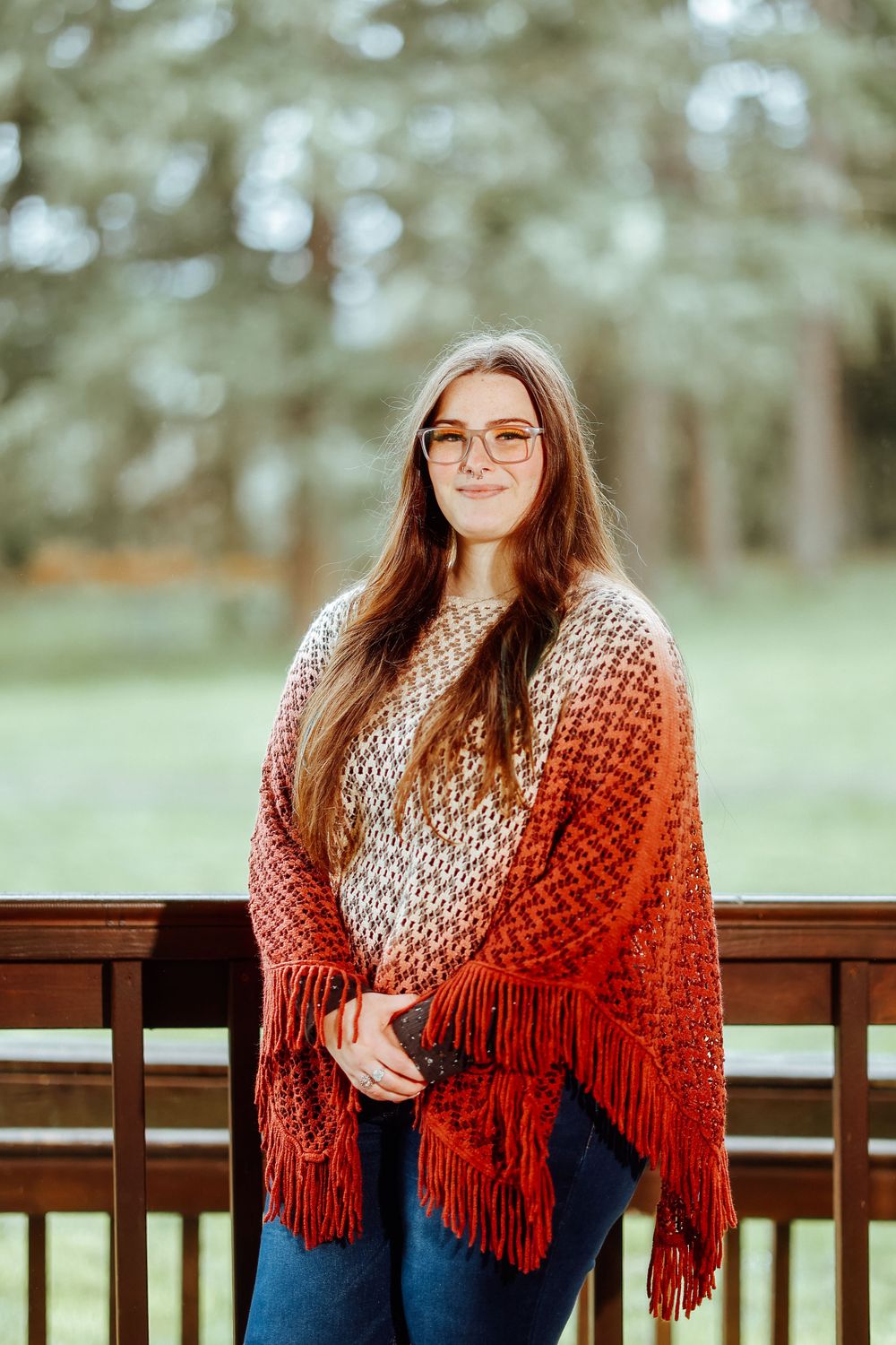 Person wearing ombre knit shawl poses on wooden deck with forest background.