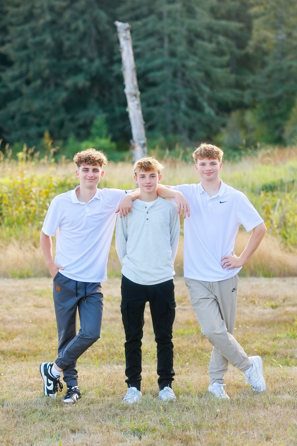Three young people in casual clothing pose together in a grassy field with evergreen trees in the background.