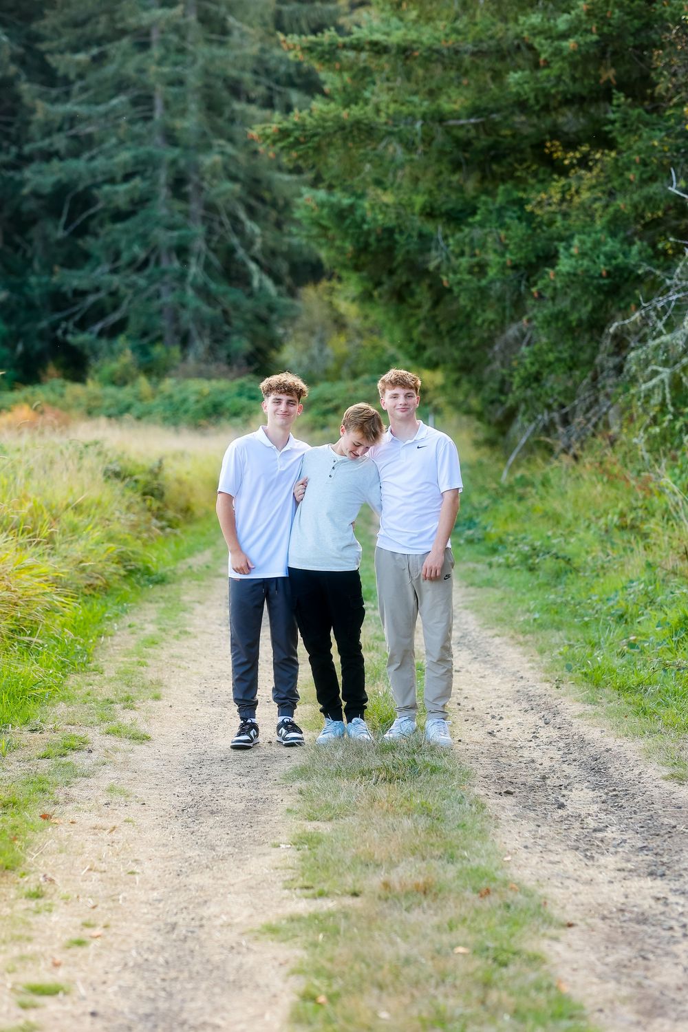Three people in light colored shirts stand together on a dirt path surrounded by evergreen trees and green foliage.