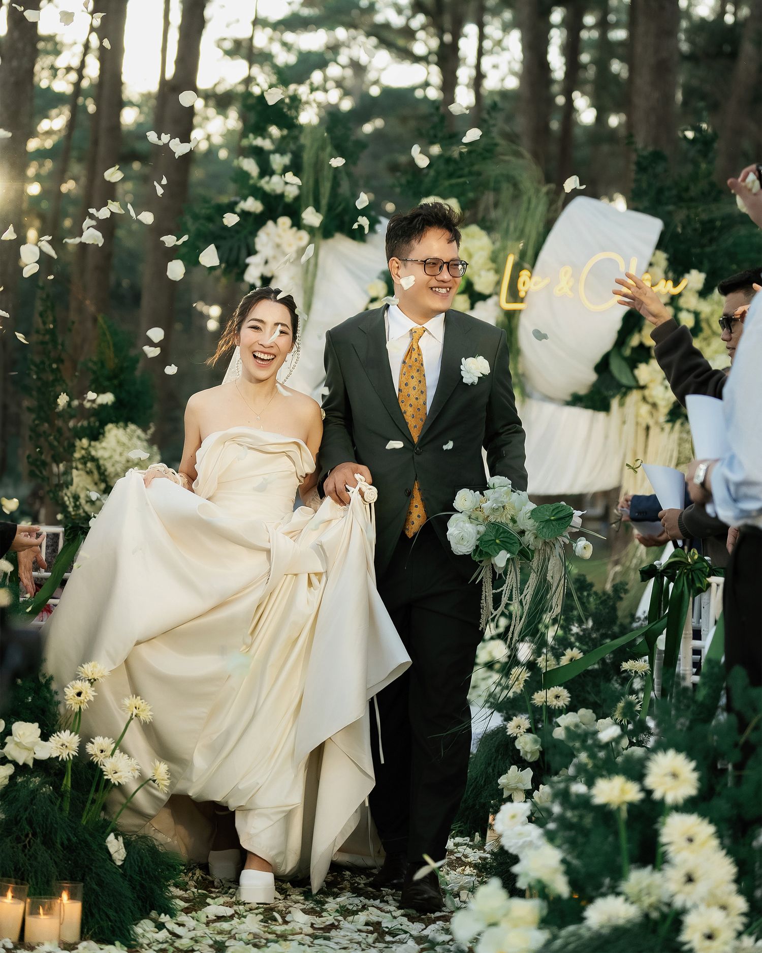 A couple walks down a forest path surrounded by flowers and greenery during an outdoor wedding celebration.