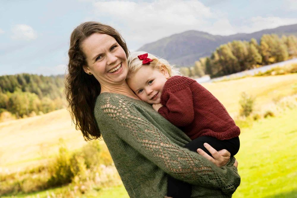 Mor som holder liten datter og smiler og ser i kamera med Lyngdal i bakgrunnen