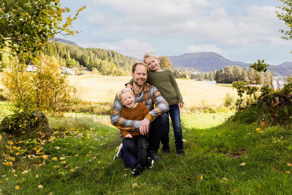 Pappa som sitter på huk med to små sønner som lener seg på han i et skogbryn i Lyngdal