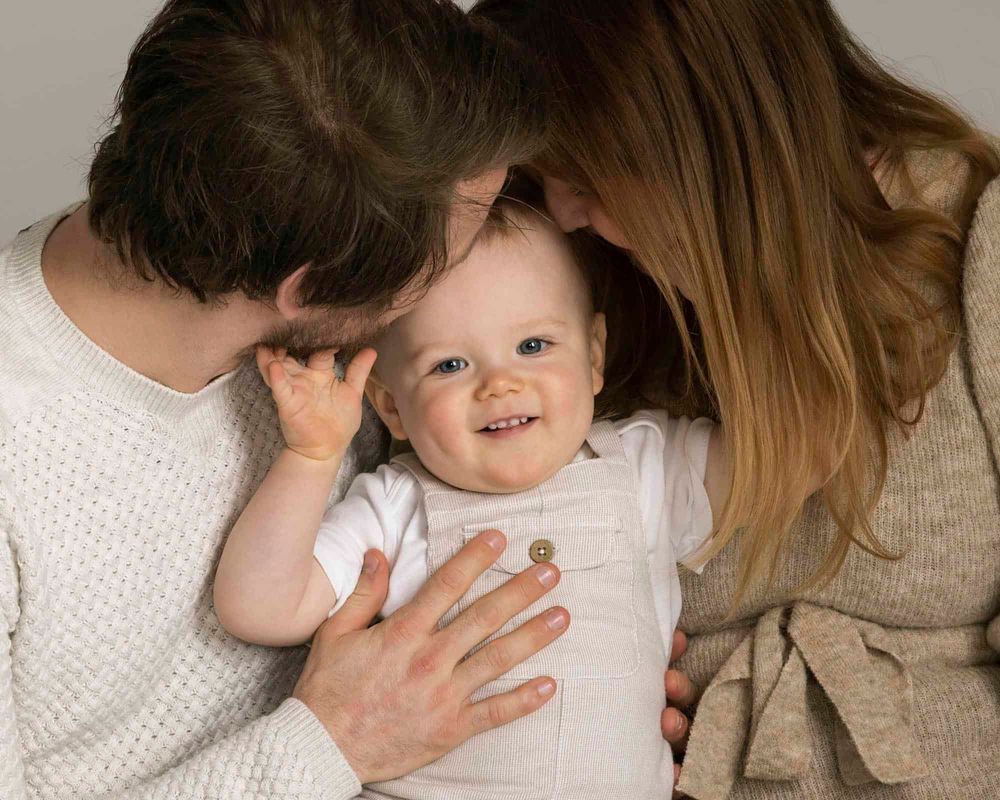 Familie med 1-åring i fotostudio i Lyngdal der gutten ser i kamera og smiler mens foreldrene kysser han på pannen