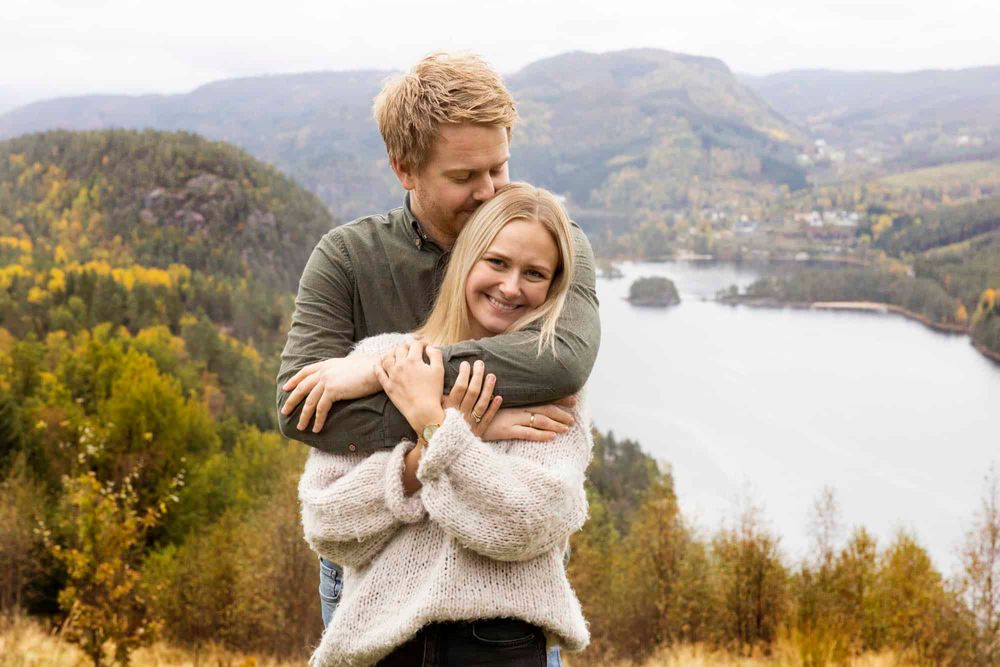 Kjærestepar på en høyde der hun smiler til kamera og han holder rundt henne bakfra og kysser henne på hodet