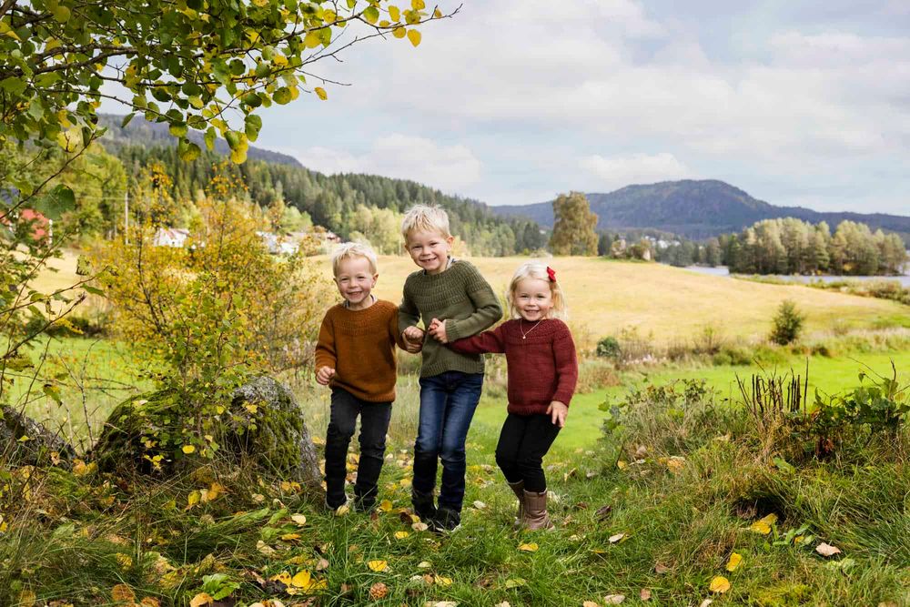 3 småsøsken som holder hender og hopper i et skogbryn i Lyngdal