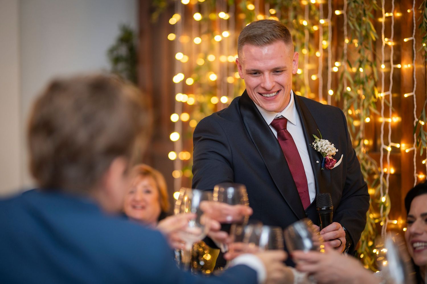 A couple shares a toast at an elegant celebration with a sparkling chandelier overhead and guests mingling in background.