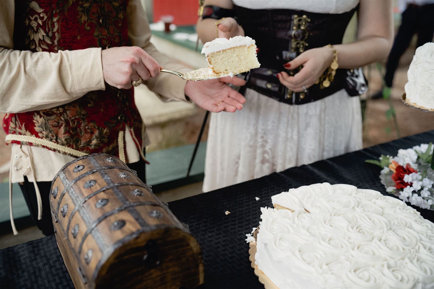 Two people serving white cake slices at a rustic table with an ornate wooden box in the foreground.
