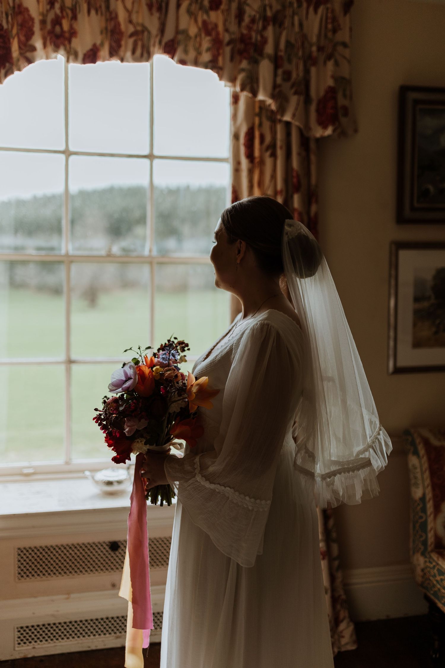 A person in a wedding dress holds a colorful bouquet while standing by a window with floral curtains at Walcott Hall.