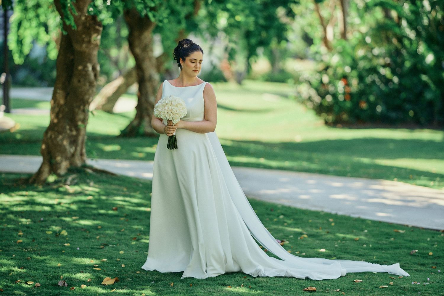 A bride in an elegant white gown with a flowing train stands on a sunlit garden path surrounded by lush greenery. Jamaican Photographer