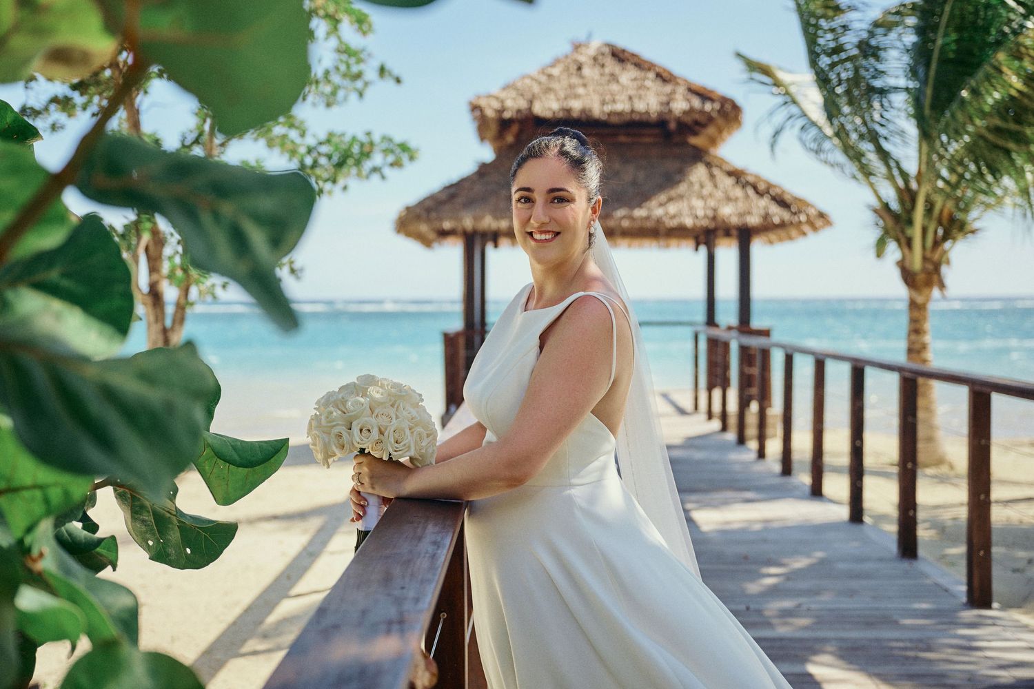 Someone in a flowing white wedding dress poses on a tropical beach boardwalk with thatched huts and palm trees. Jamaican Photographer