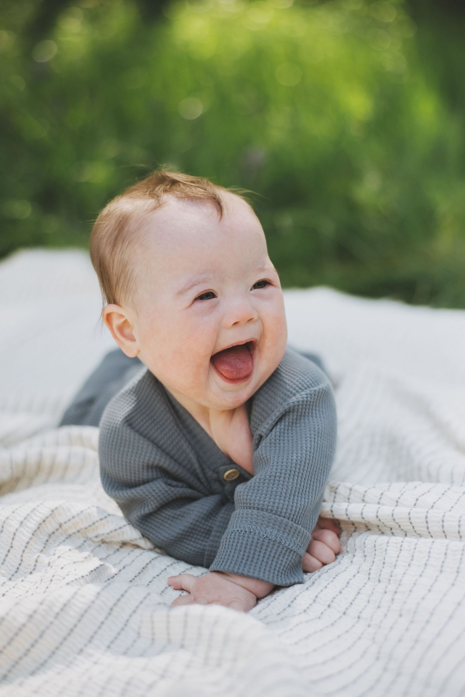 Happy baby wearing gray cardigan lies on white knitted blanket outdoors with natural green background.