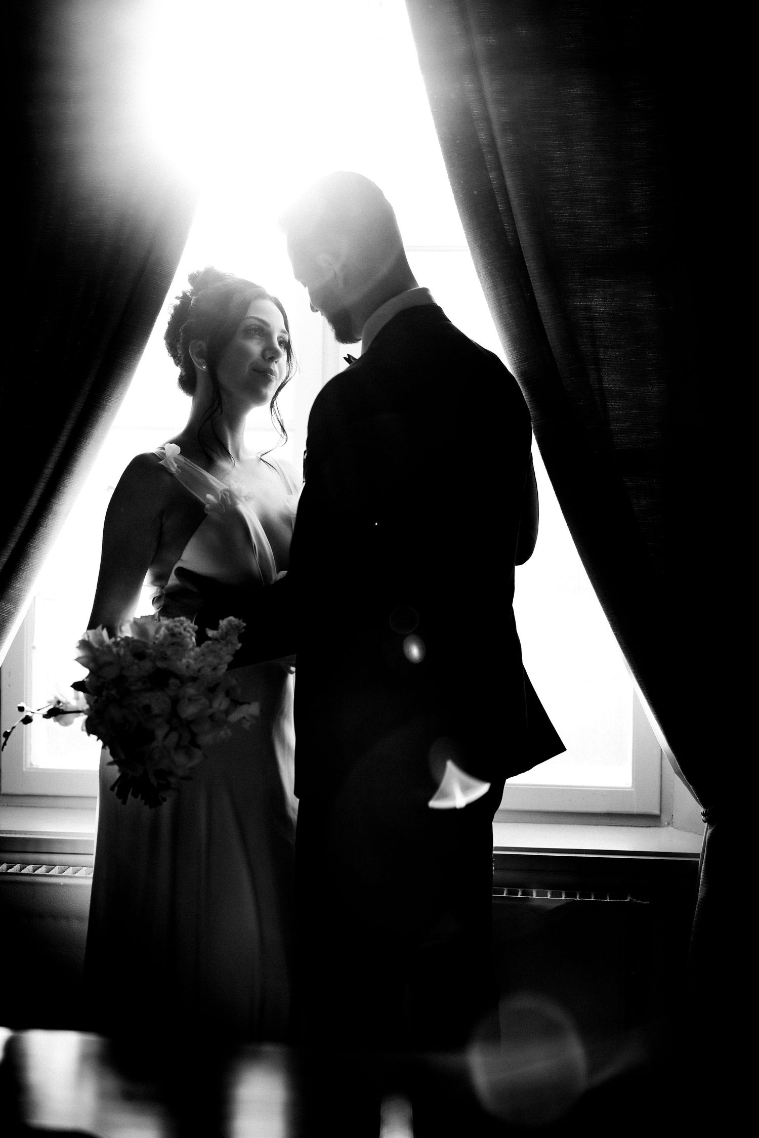 Dramatic black and white wedding silhouette of couple by window with bride holding bouquet and light streaming behind them.