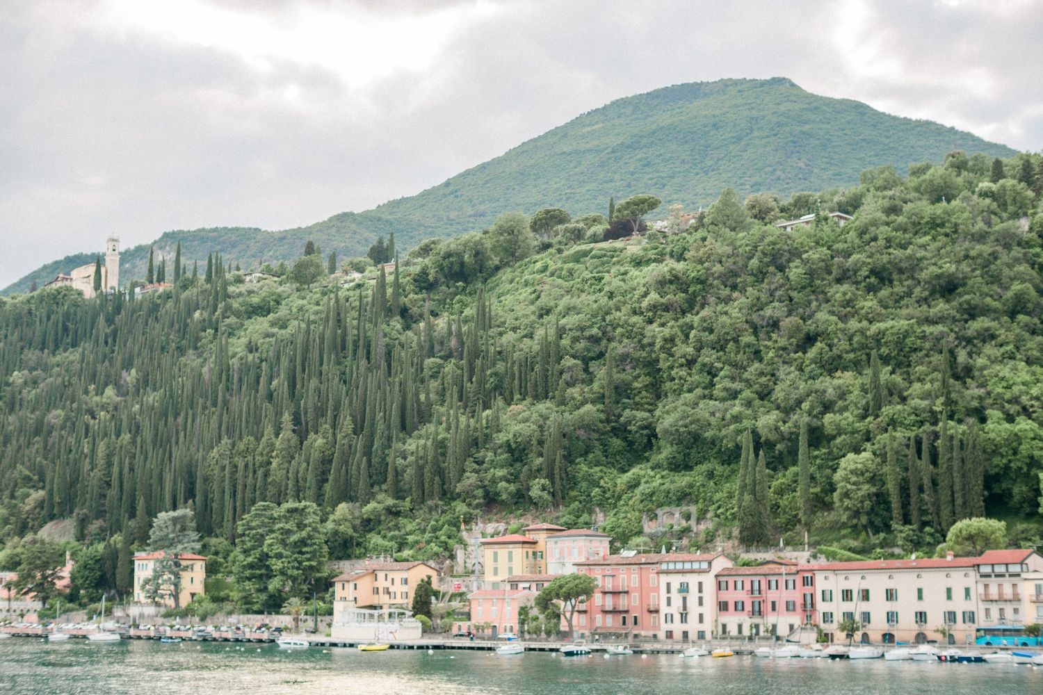 Italian coastal village with colourful buildings nestled against green hillside covered in cypress trees above a bay in lake Garda